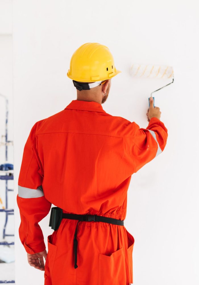 Young foreman standing from back in orange work clothes and yellow hardhat using painting roller in new apartment at work